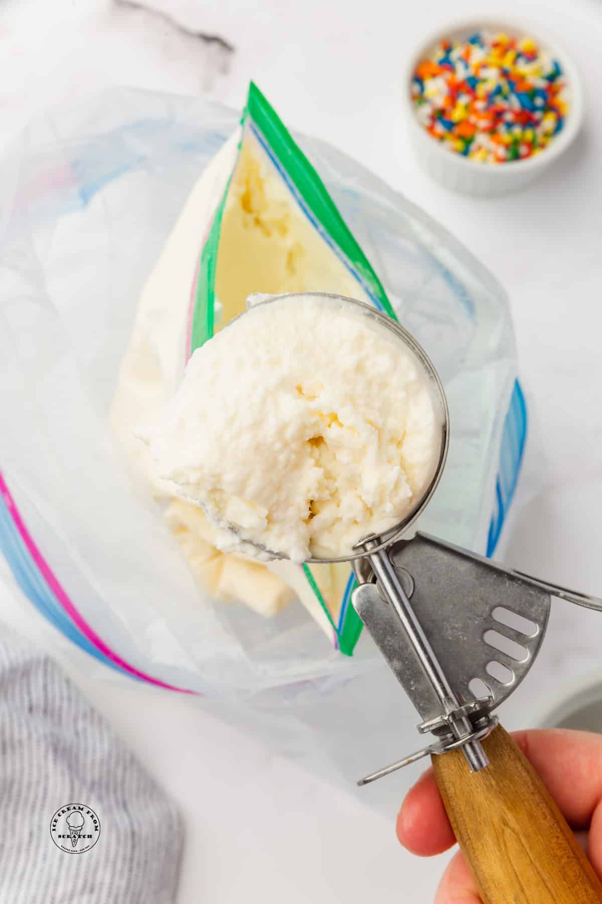 Vanilla ice cream in a metal ice cream scoop, above a double zip log bag ice cream making set up. A bowl of rainbow sprinkles is in the background.