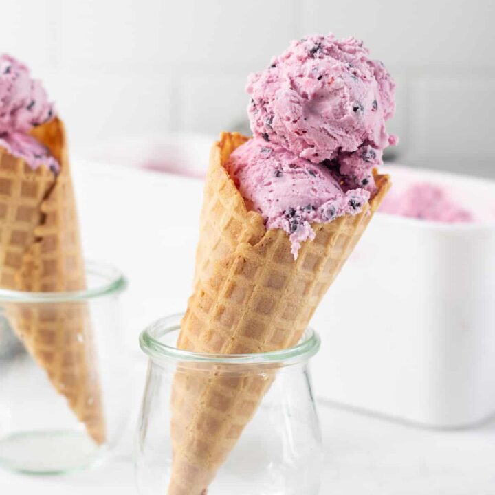 Two small jars holding up waffle cones filled with purple huckleberry ice cream.