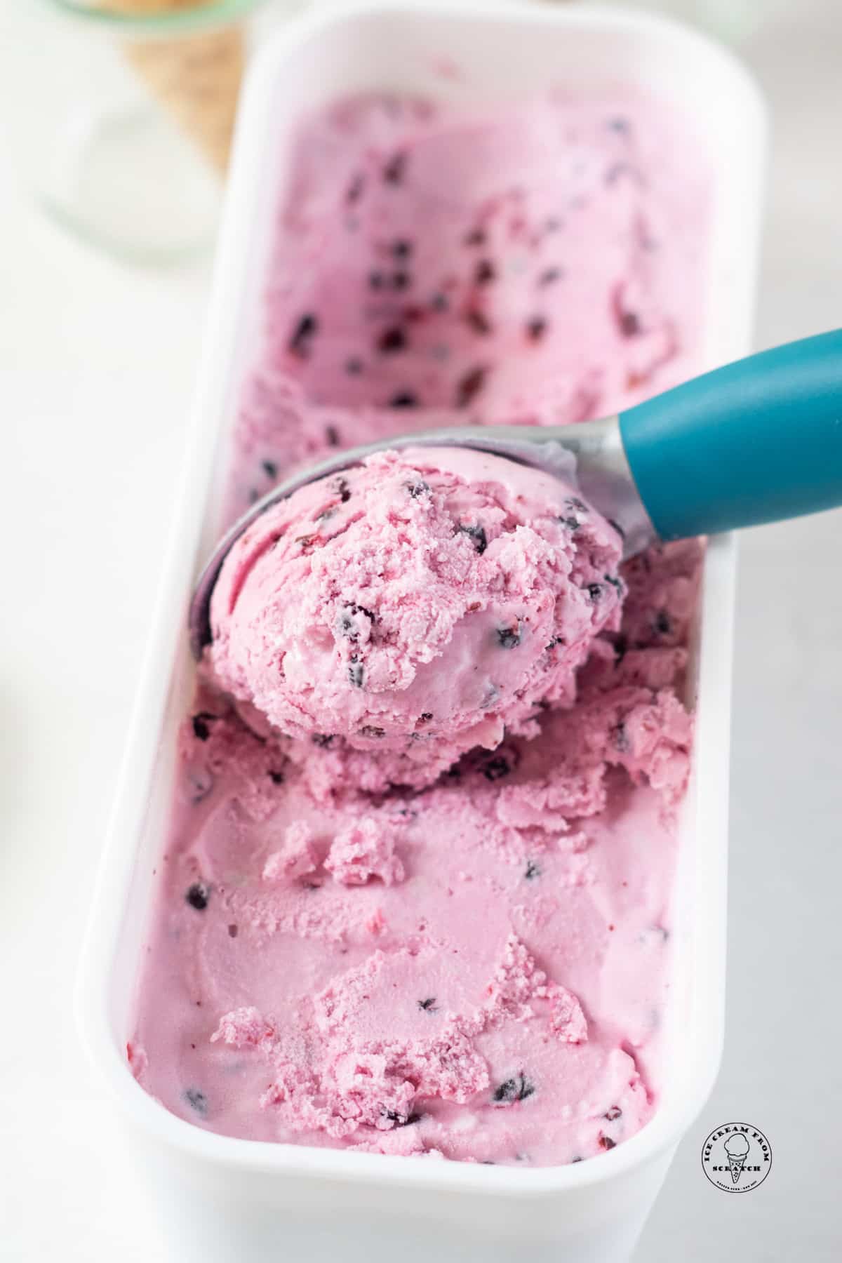 a blue ice cream scoop scooping huckleberry ice cream from a white plastic container.