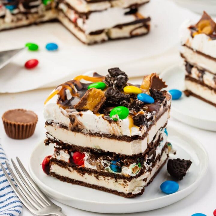 a square piece of ice cream cake made with ice cream sandwiches layered with cool whip, topped with m&Ms, reeses cups, and oreo pieces.