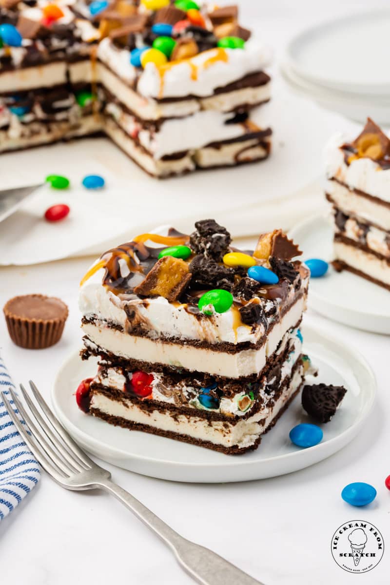 a square piece of ice cream cake made with ice cream sandwiches layered with cool whip, topped with m&Ms, reeses cups, and oreo pieces.