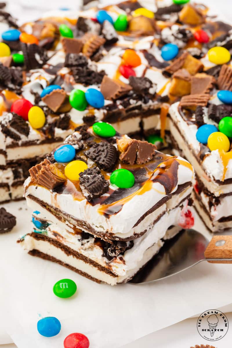 a square piece of ice cream sandwich cake being removed from the rest of the cake with a spatula. The slice has layers of ice cream sandwiches and cool whip, and is topped with candy and oreo pieces.