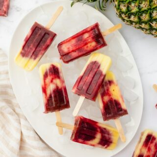 8 swirled cherry pineapple popsicles on a tray of ice, viewed from above. The tray is next to a whole fresh pineapple.
