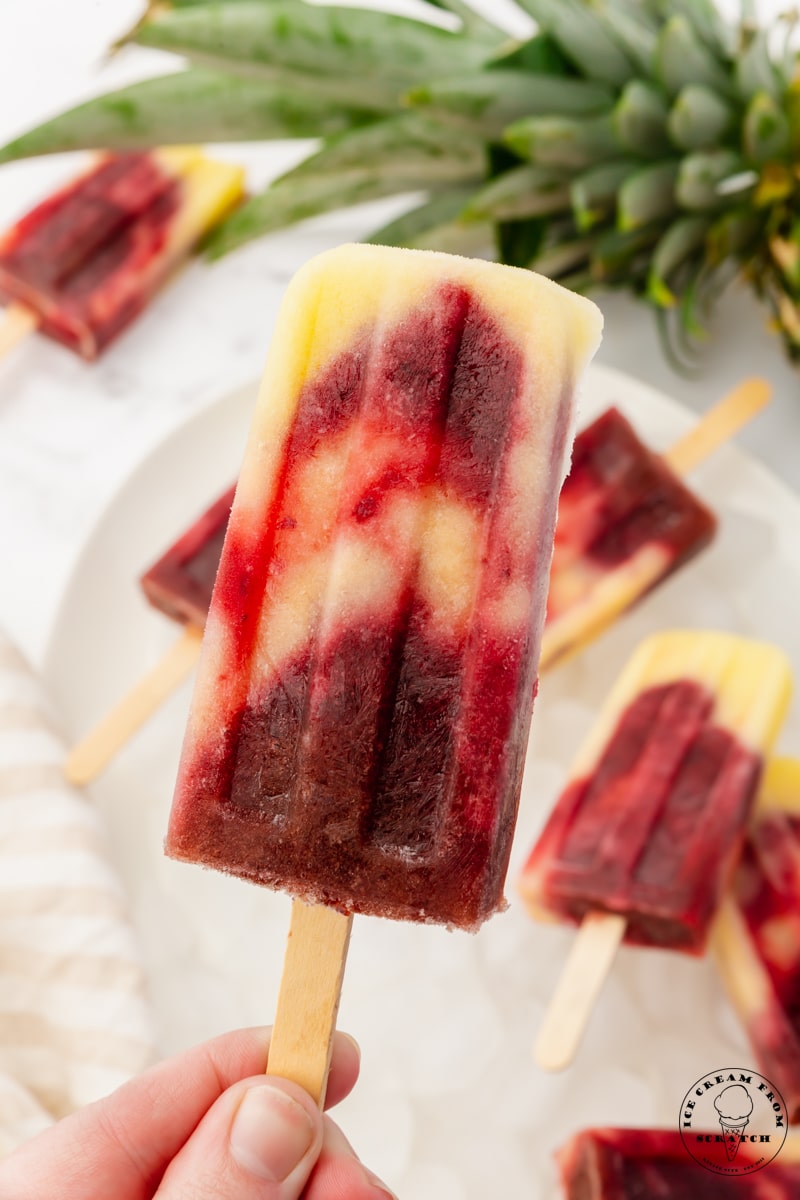 a hand holding a homemade cherry pineapple popsicle up by the stick. In the background are more popsicles and a fresh pineapple top.