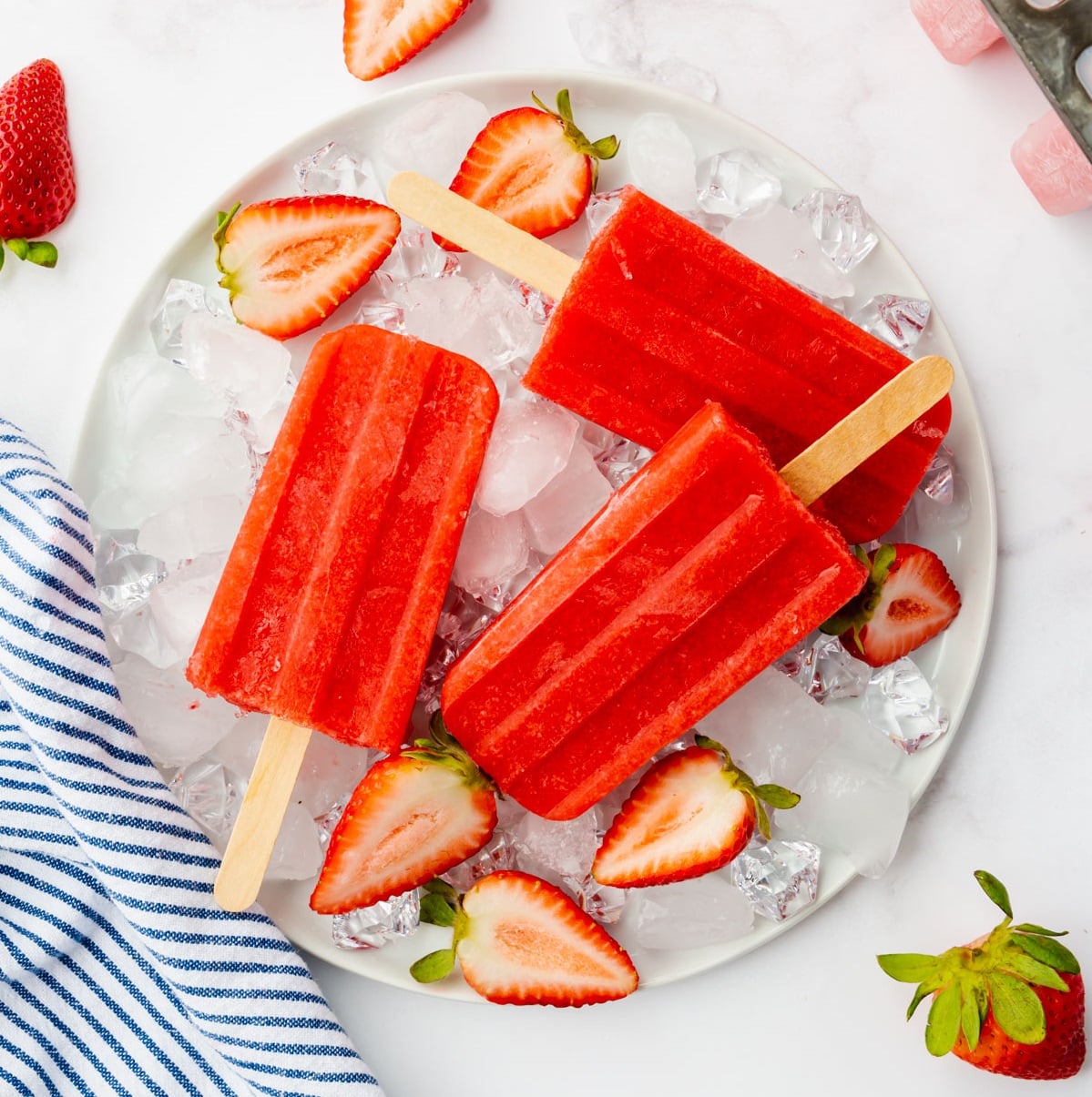 A round plate of ice topped with three homemade strawberry popsicles and halved berries.