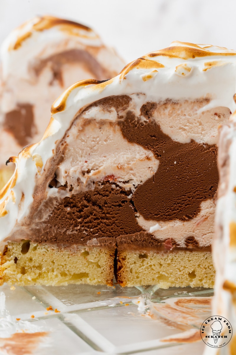 Closeup view of the inside of a slice of baked Alaska. There is a bottom layer of cake, topped with chocolate and strawberry ice cream swirls, then topped with fluffy meringue.