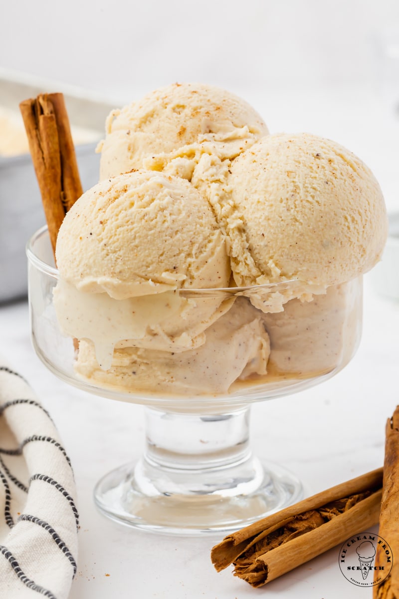 A footed glass dish holding 5 scoops of homemade eggnog ice cream, speckled with cinnamon and spices. There are rolled cinnamon sticks on the counter next to the dish, and stuck into the ice cream.