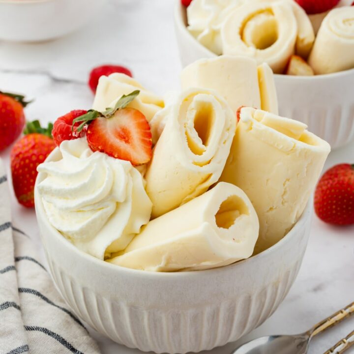 a white bowl filled with rolls of vanilla ice cream, topped with whipped cream and fresh berries. There are two spoons next to the bowl.