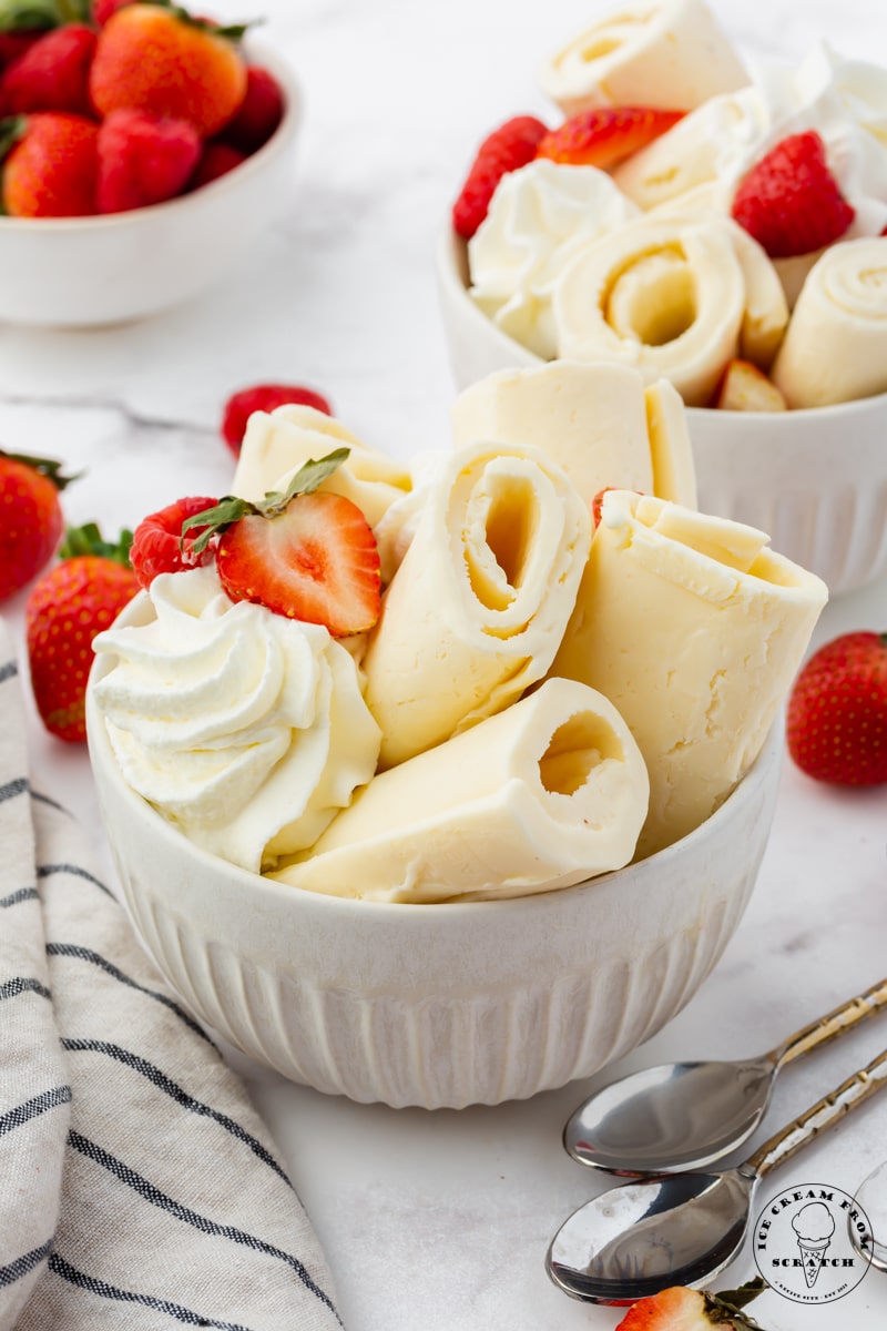 a white bowl filled with rolls of vanilla ice cream, topped with whipped cream and fresh berries. There are two spoons next to the bowl.