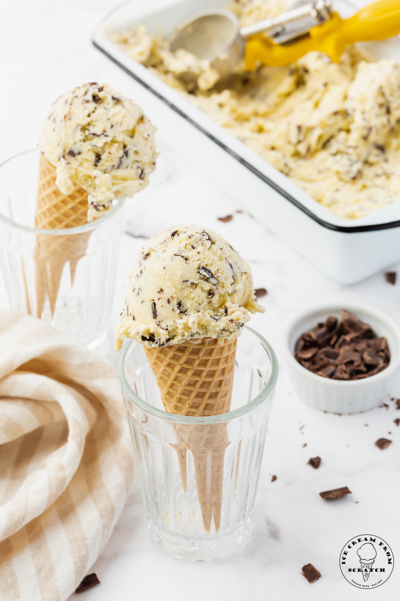 Two cones of homemade stracciatella ice cream held up in glasses, next to a white loaf pan filled with ice cream.