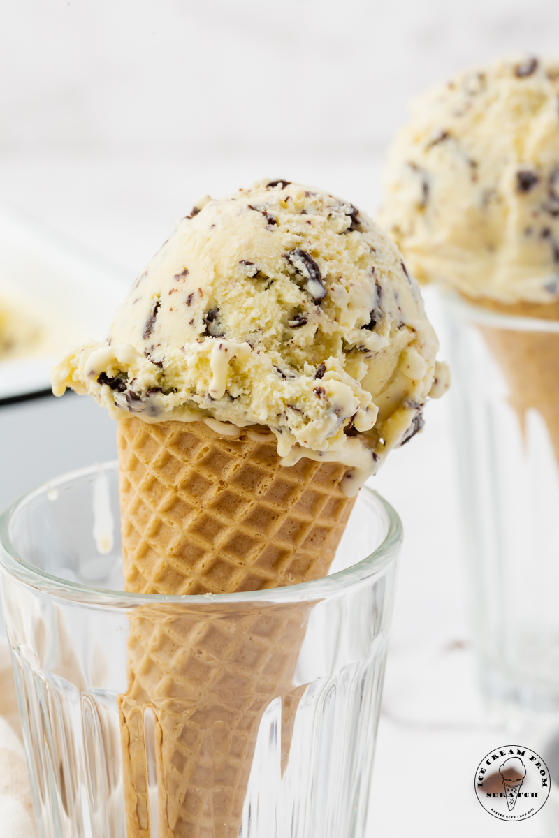 a scoop of stracciatella ice cream with shards of chocolate on top of a sugar cone. The cone is held upright in a glass.