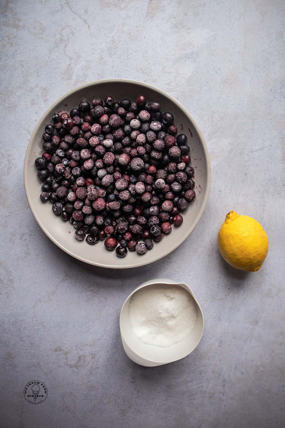 A bowl of frozen blueberries next to a small bowl of sugar and a fresh lemon