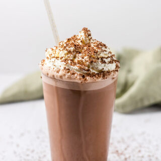 A pint glass holding a coffee milkshake, topped with fresh whipped cream and chocolate shavings. There is a clear straw in the glass.