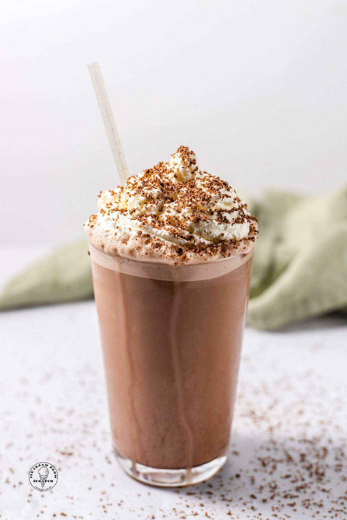 A pint glass holding a coffee milkshake, topped with fresh whipped cream and chocolate shavings. There is a clear straw in the glass.