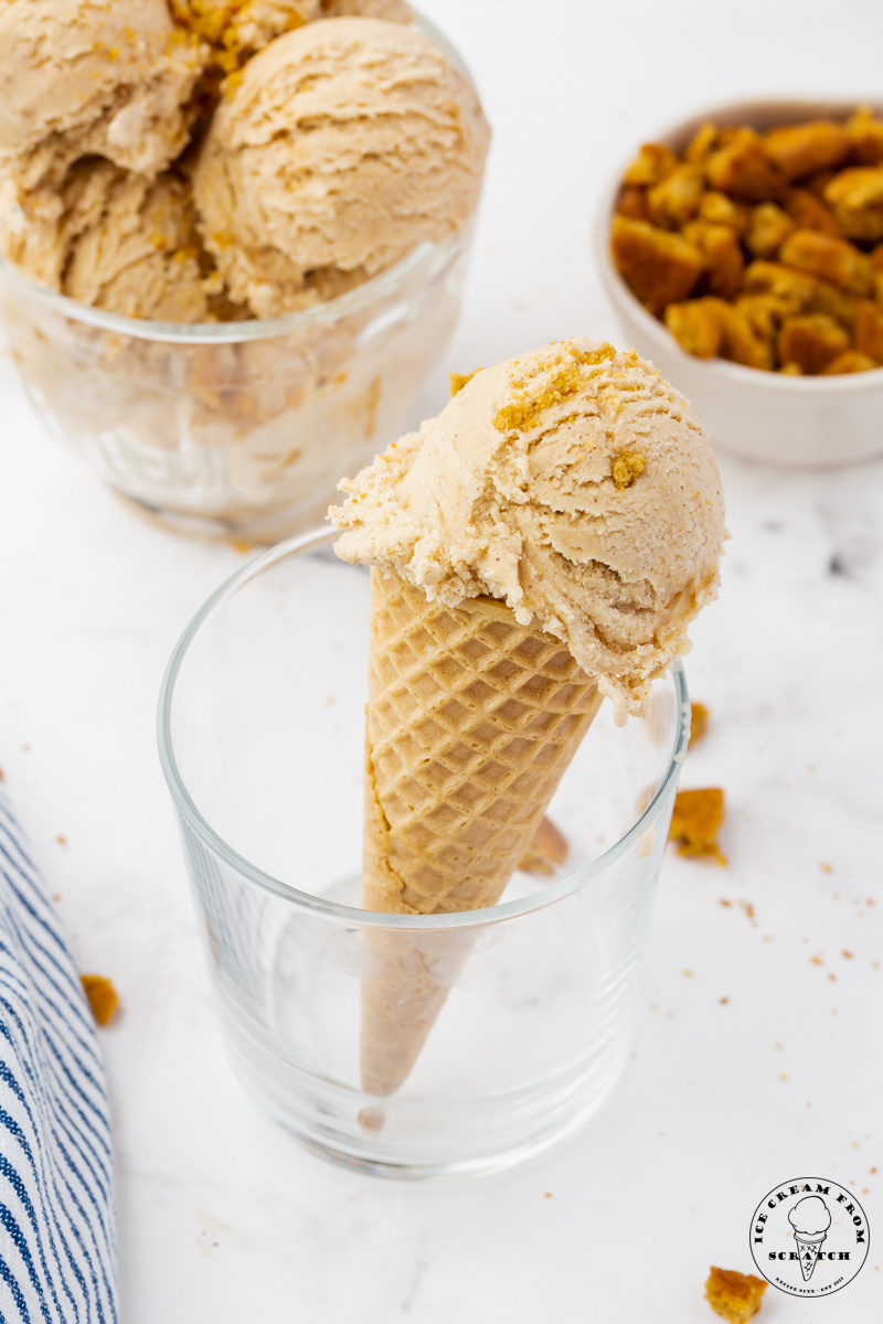 a sugar cone topped with a scoop of cooked butter ice cream. the cone is held up in a glass. Behind this is a glass bowl of ice cream and a bowl of crushed speculoos cookies.