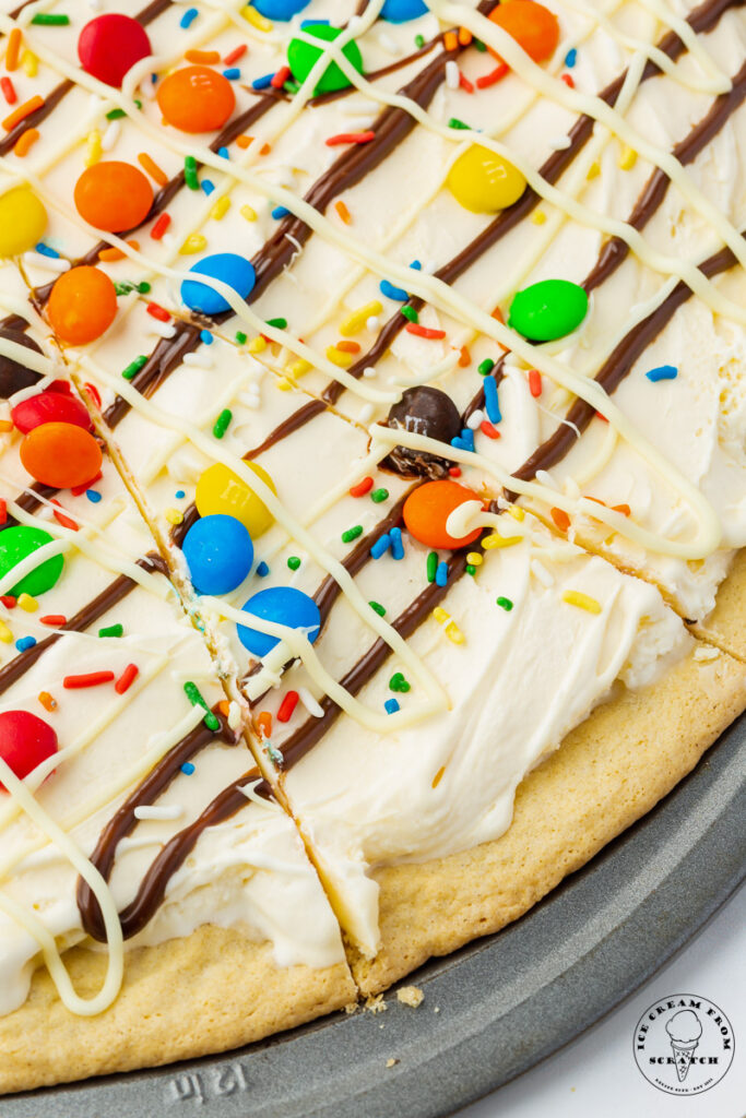 closeup of the edge of a homemade ice cream pizza in a metal pizza pan. the dessert is topped with white chocolate, hot fudge, sprinkles, and m&ms