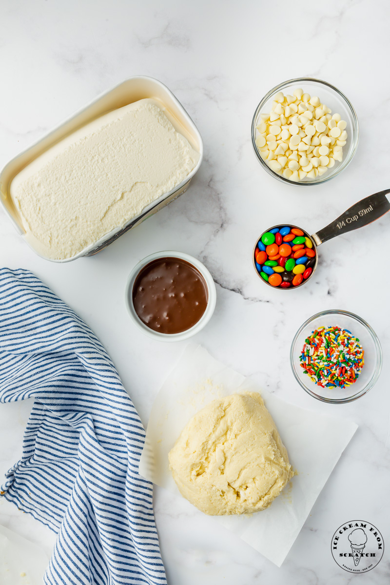 The ingredients needed to make ice cream pizza, including sugar cookie dough, fudge, white chocolate chips, and sprinkles.
