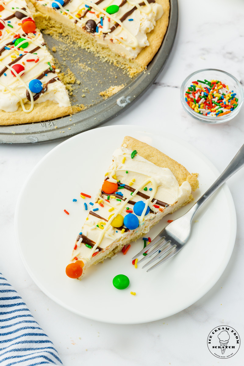 a white plate holding a slice of ice cream pizza and a fork. behind the plate is a small bowl of rainbow sprinkles and the remaining cookie ice cream pizza on a pizza pan.