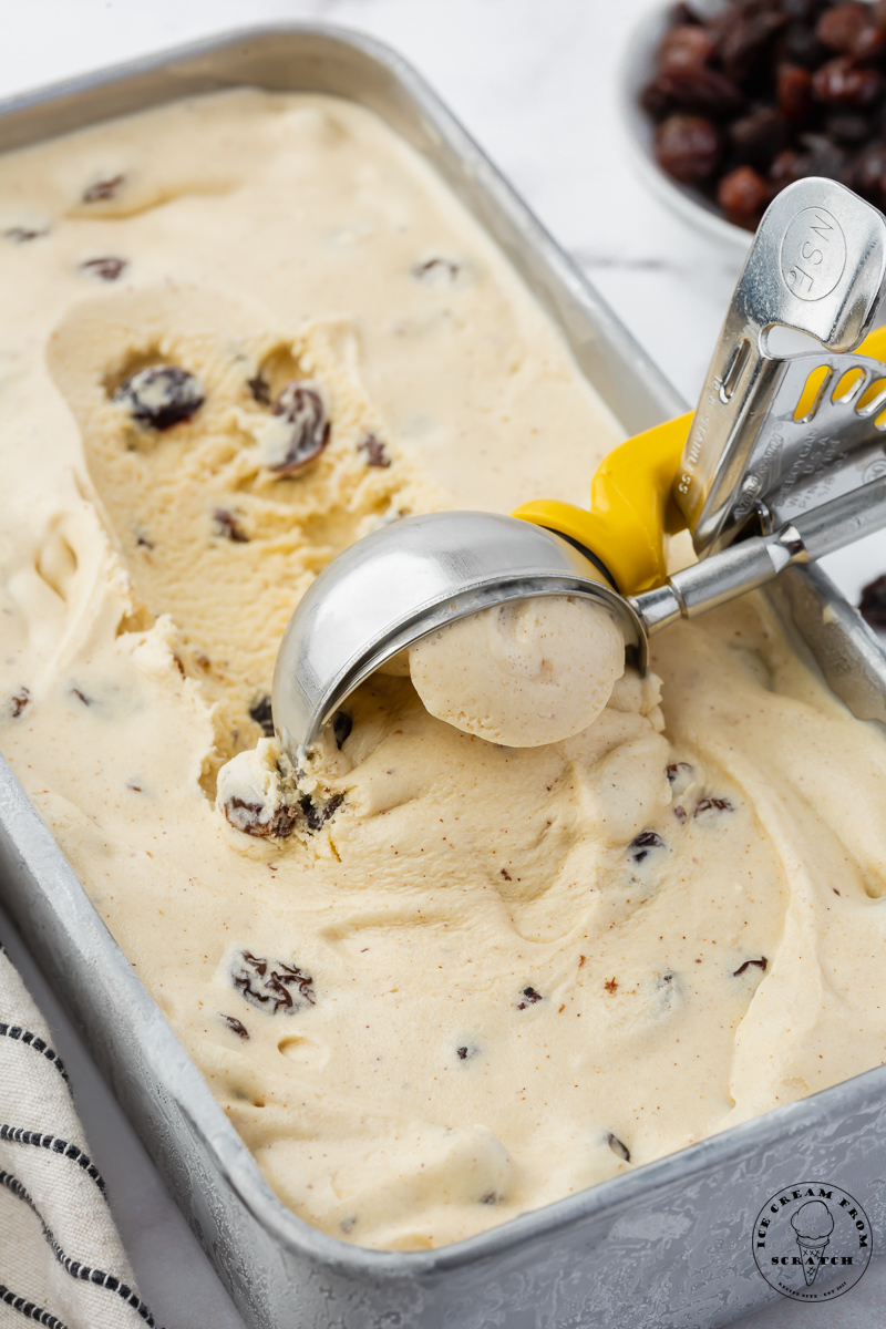 a metal loaf pan filled with homemade rum raisin ice cream. a metal ice cream scoop is pulling up a scoop of the ice cream.