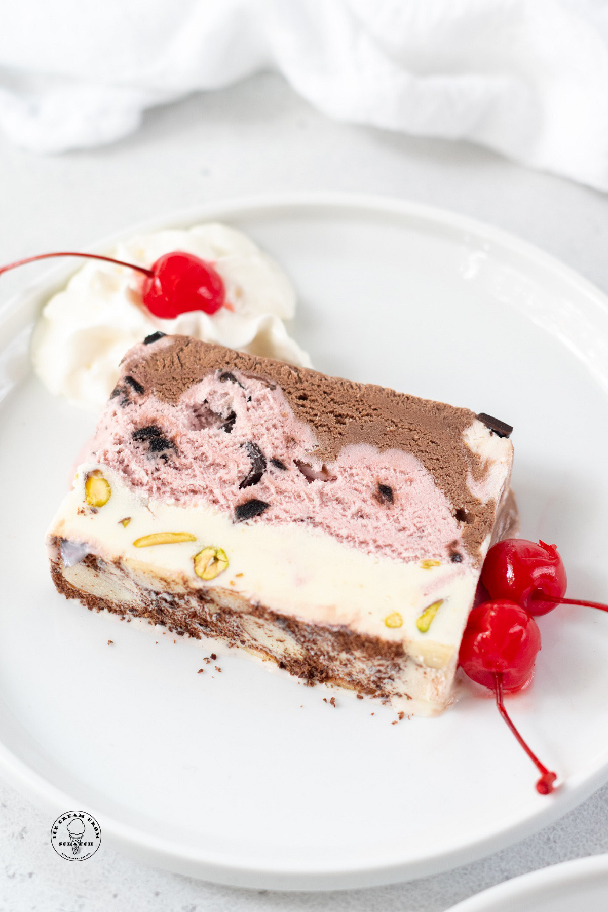 a slice of spumoni with layers of pistacio, cherry, and chocolate ice cream, on a plate with maraschino cherry garnish.