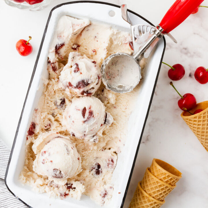 A white loaf pan filled with cherry gelato. A red and metal ice cream scoop is in the pan, and the ice cream has been scooped. Around the pan are fresh cherries and sugar cones.