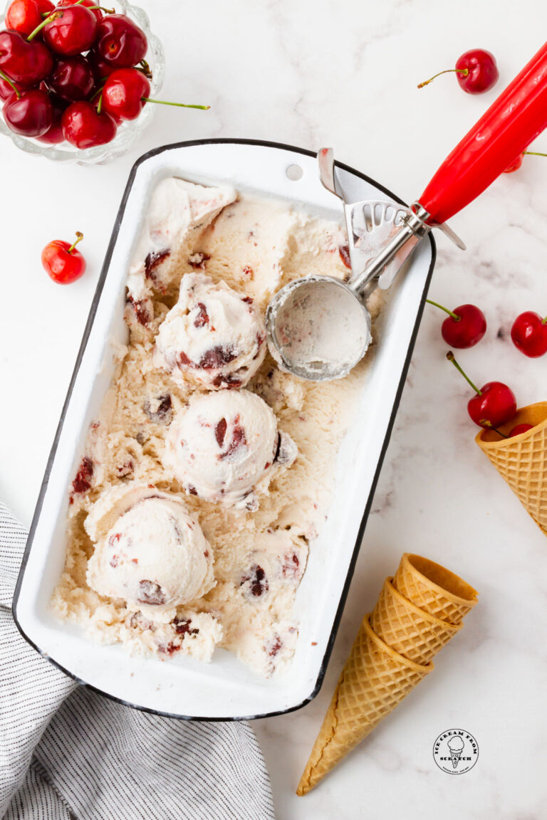 A white loaf pan filled with cherry gelato. A red and metal ice cream scoop is in the pan, and the ice cream has been scooped. Around the pan are fresh cherries and sugar cones.