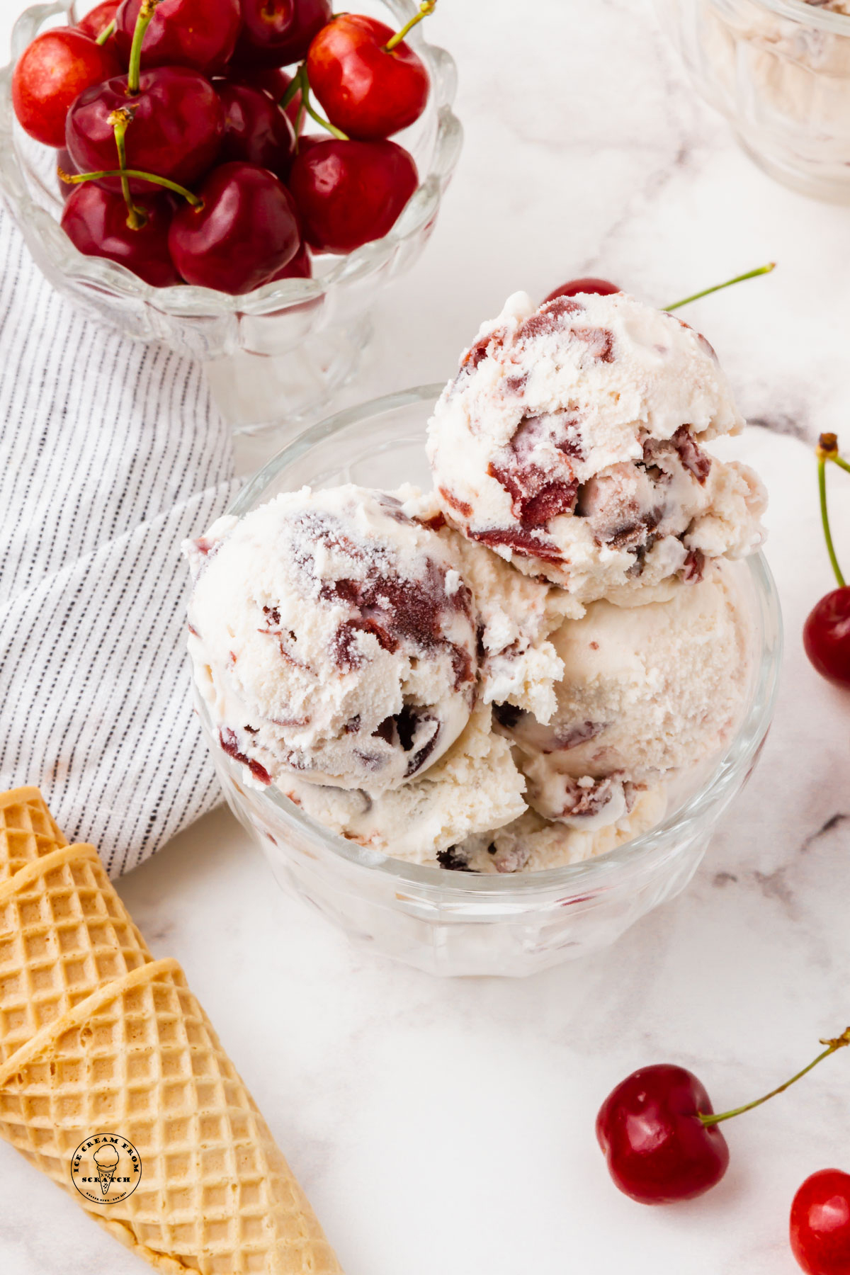 a glass bowl filled with scoops of cherry gelato, next to a bowl of fresh cherries and sugar cones.