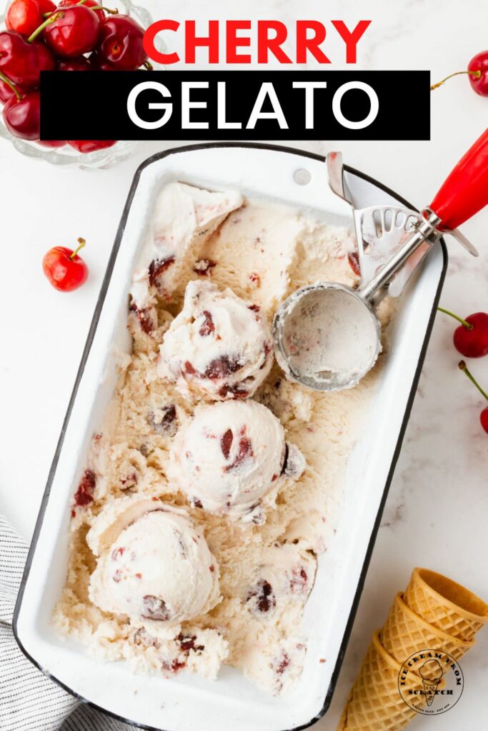 A white loaf pan filled with cherry gelato. A red and metal ice cream scoop is in the pan, and the ice cream has been scooped. Around the pan are fresh cherries and sugar cones.