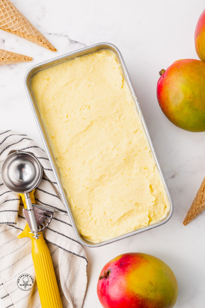 a loaf pan filled with creamy mango gelato. Next to the pan are fresh mangoes and a yellow metal ice cream scoop.