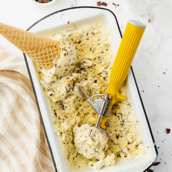 stracciatella gelato in a white loaf pan. A metal scoop with a yellow handle is making scoops for a cone.