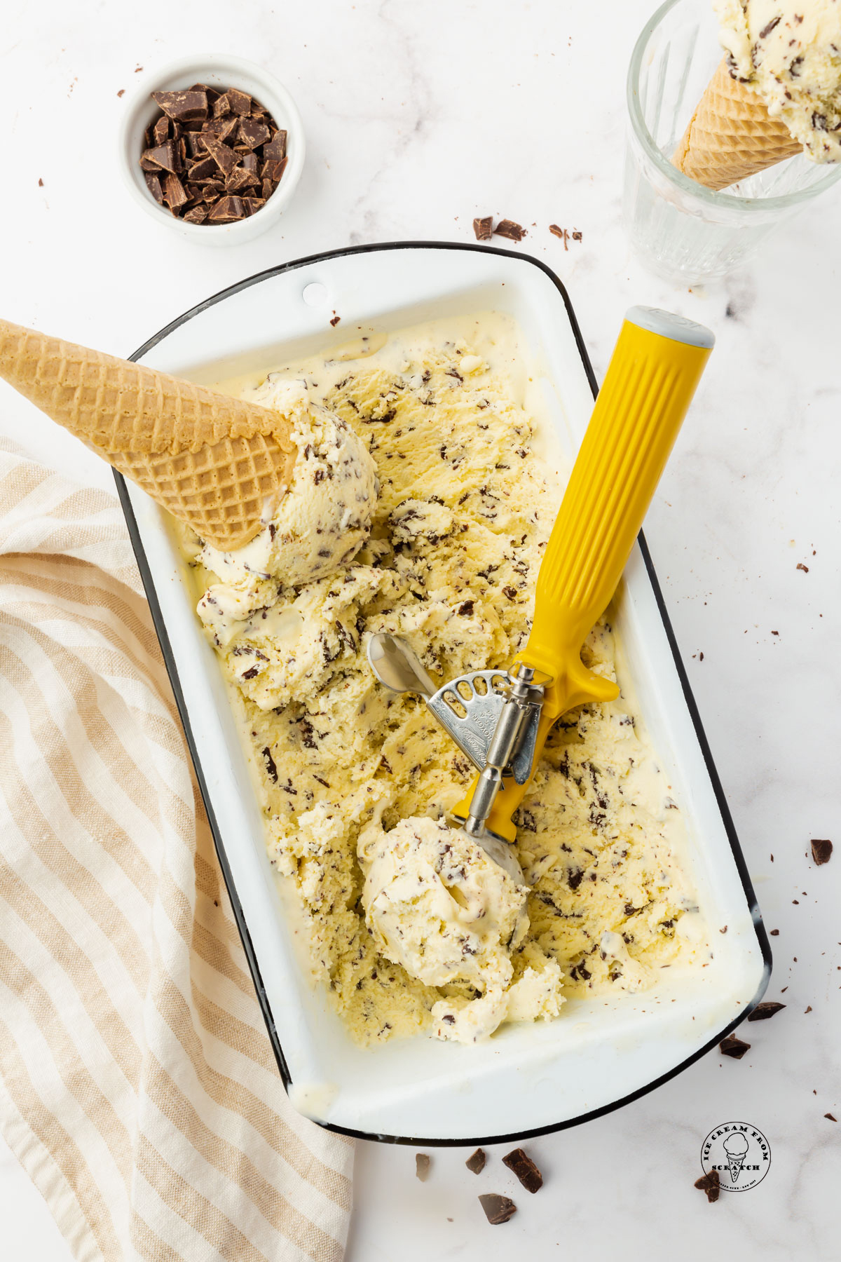 stracciatella gelato in a white loaf pan. A metal scoop with a yellow handle is making scoops for a cone.