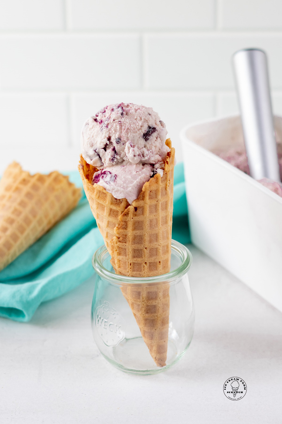 Pink amaretto ice cream with cherry chunks, in a waffle cone, propped up in a small glass jar.