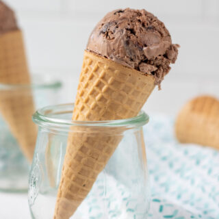 a sugar cone topped with one scoop of chocolate ice cream, held upright in a small jelly jar.