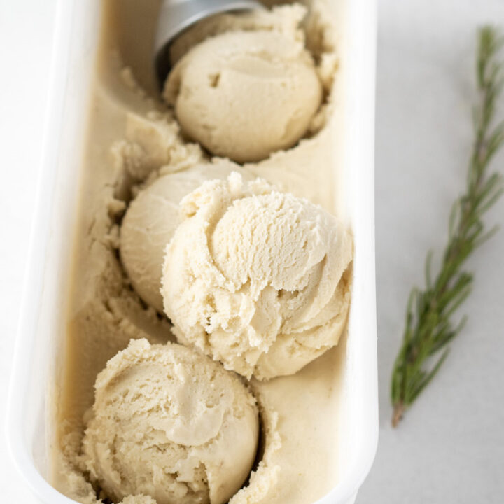 scoops of homemade rosemary ice cream in a white plastic rectangular container with a metal ice cream scoop.