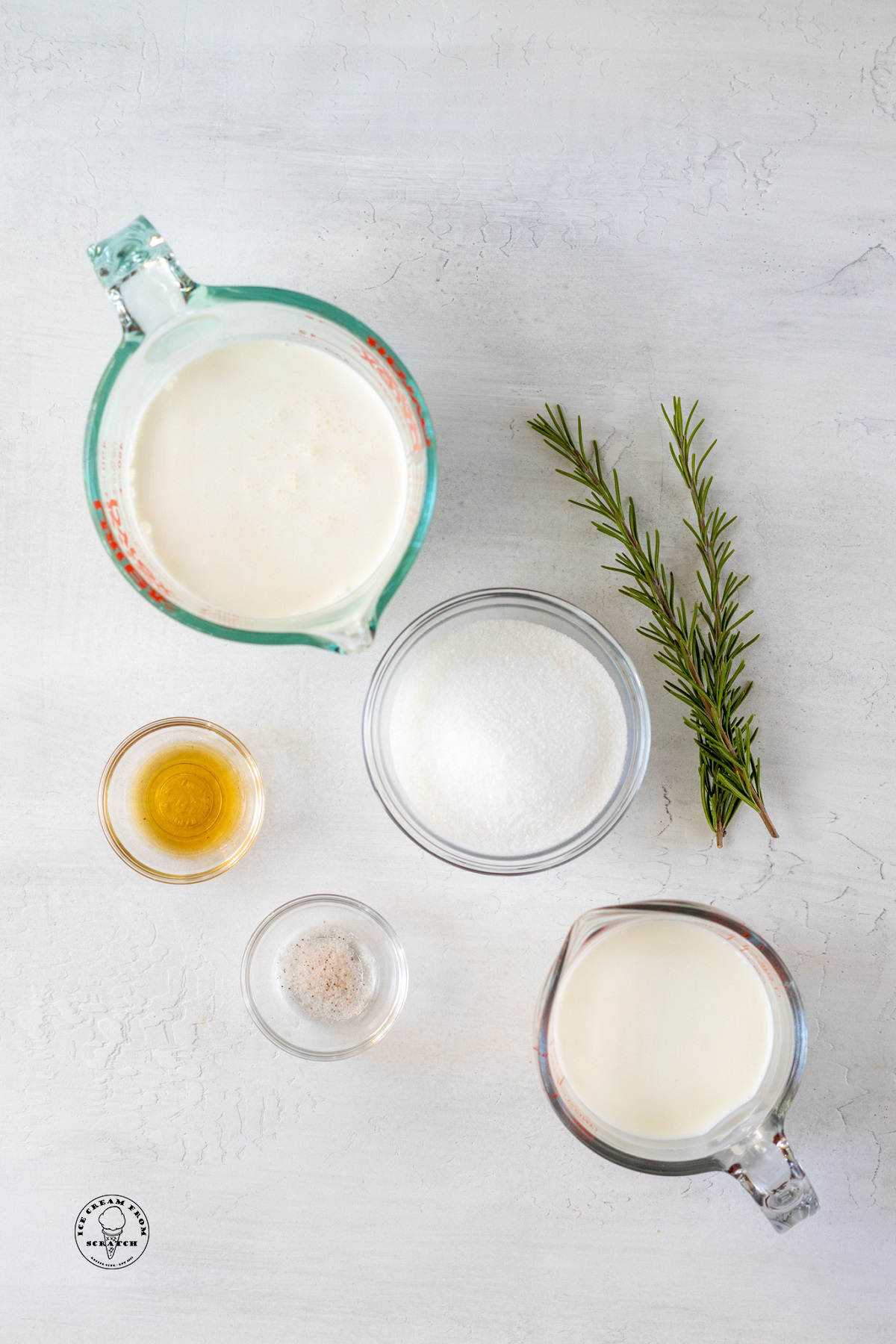 Milk, cream, sugar, salt, and vanilla, measured into glass bowls and measuring cups, next to two sprigs of rosemary on a stone counter.