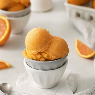 two small bowls stacked, holding three scoops of orange sherbet. In the background are fresh oranges.