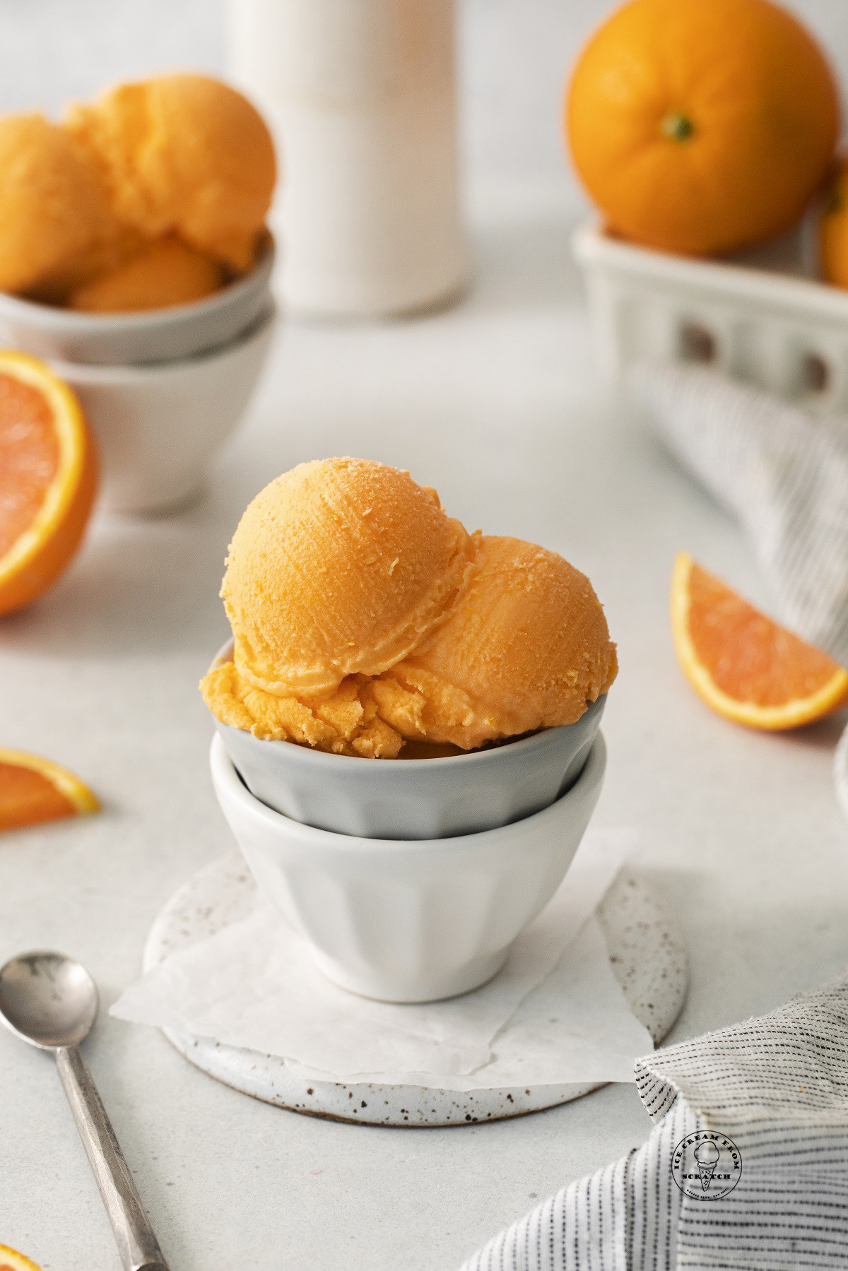 two small bowls stacked, holding three scoops of orange sherbet. In the background are fresh oranges.