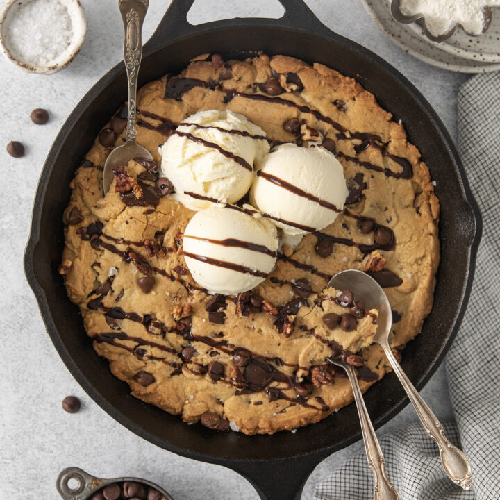 A giant cookie (pazookie) in a cast iron pan topped with scoops of ice cream. Three spoons are digging in.