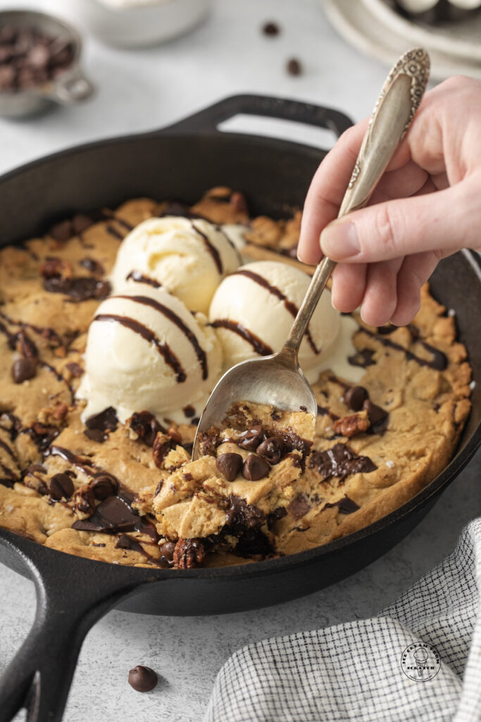 a hand holding a large spoon and eating directly from a cast iron pan holding a pazookie and ice cream.