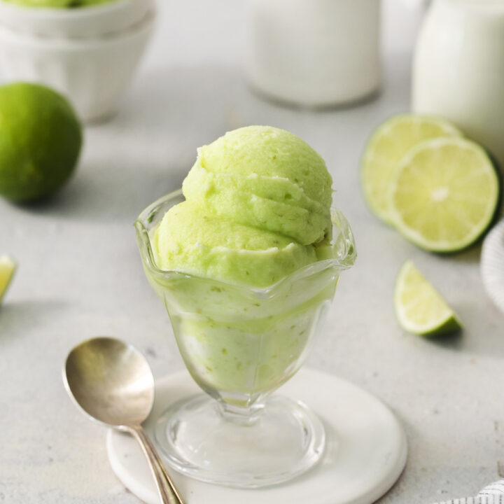 a footed glass ice cream dish filled with scoops of homemade lime sherbet. There is a silver spoon next to the dish and sliced limes in the background.