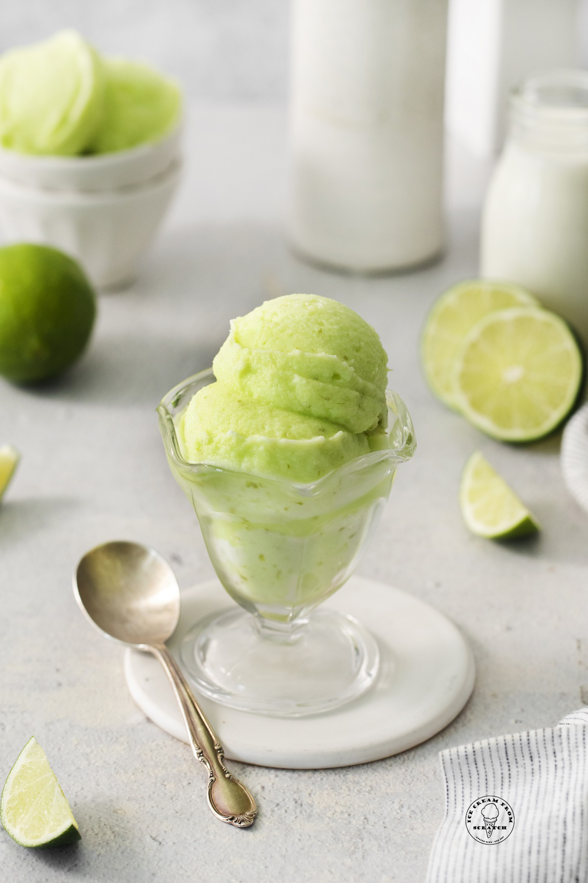 a footed glass ice cream dish filled with scoops of homemade lime sherbet. There is a silver spoon next to the dish and sliced limes in the background.