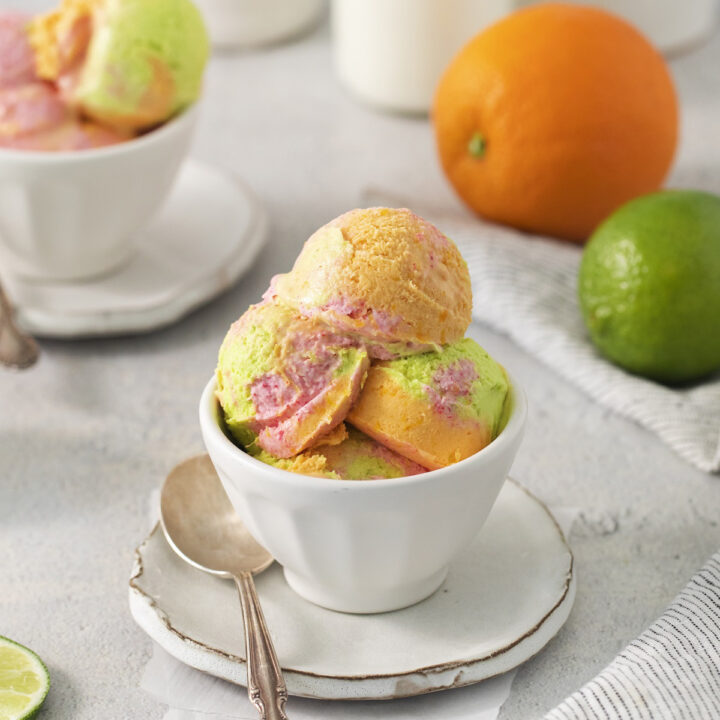 A bowl of scoops of homemade rainbow sherbet in front of bottles of cream, an orange, and a lime.