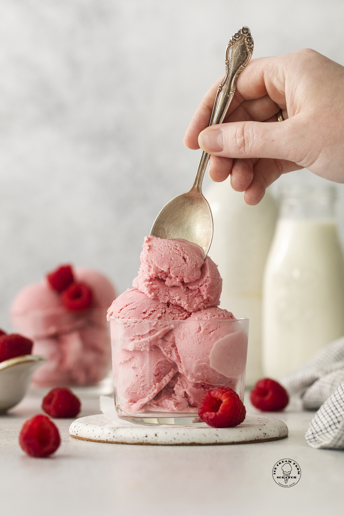 a hand holding a silver spoon that is scooping into a glass bowl filled with scoops of pink sherbet. Raspberries are next to the bowl, and bottles of cream are in the background.
