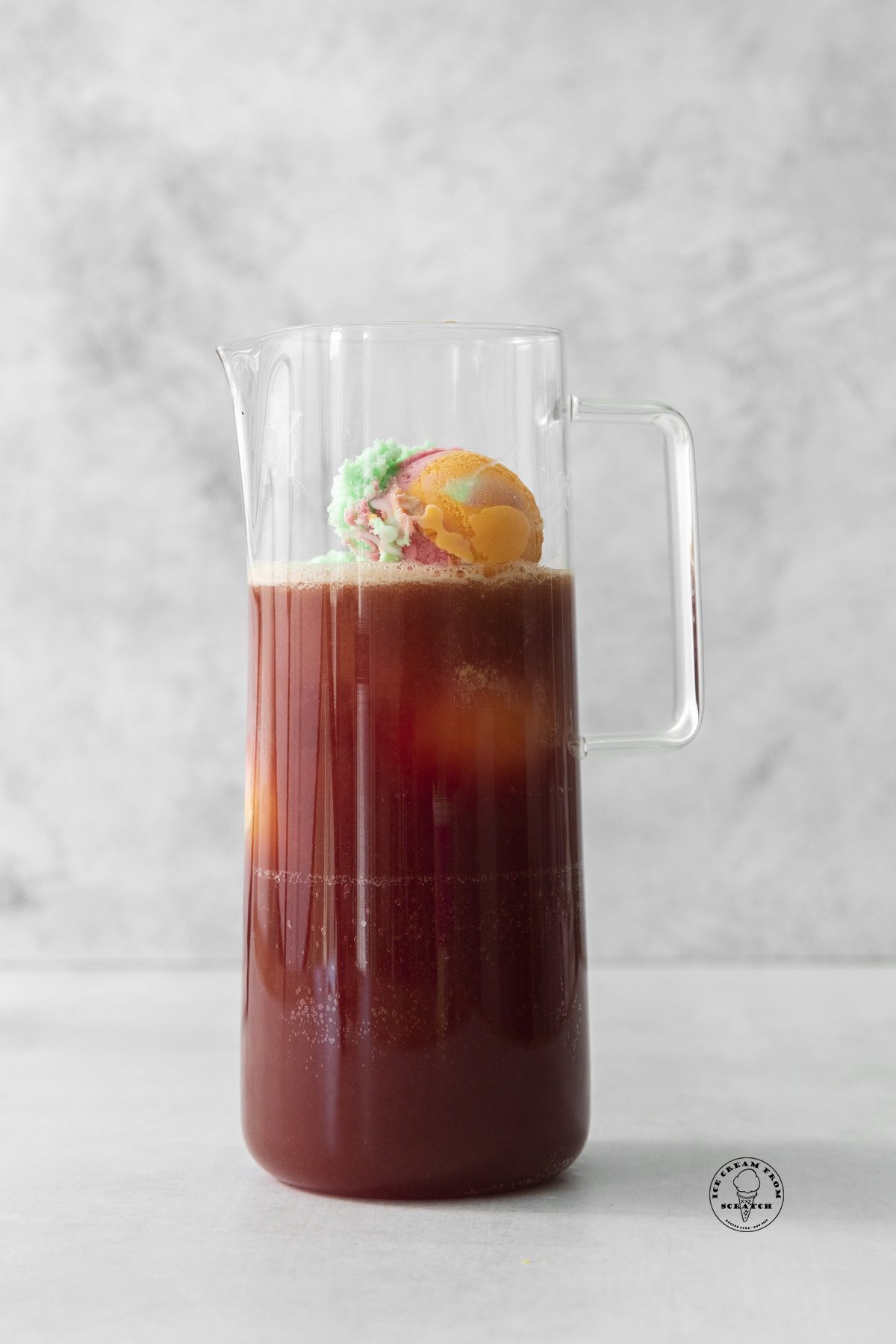 a slim clear glass pitcher filled with dark red punch and rainbow sherbet balls.