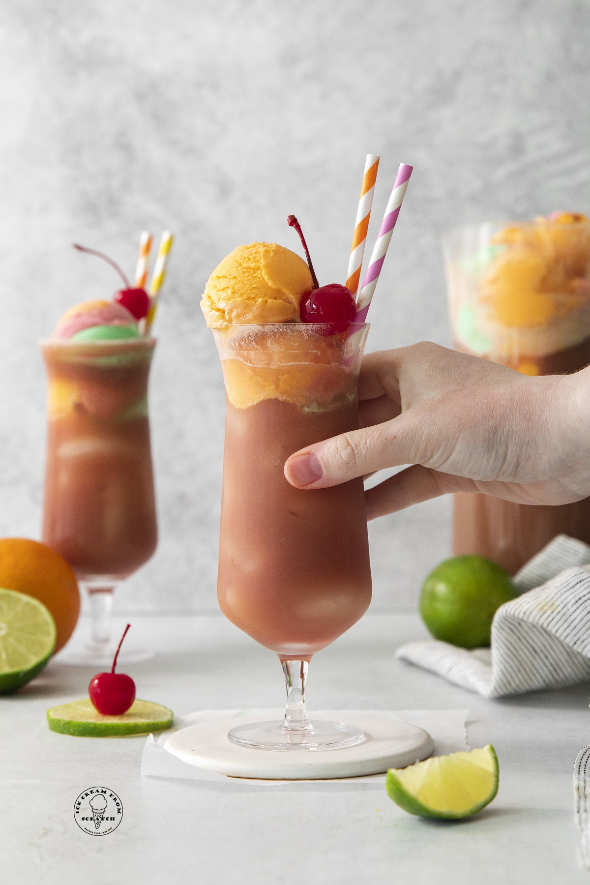 a hurricane glass filled with punch, topped with orange sherbet and a cherry.  There are two paper straws in the cup, and a hand lifts it off a white coaster.