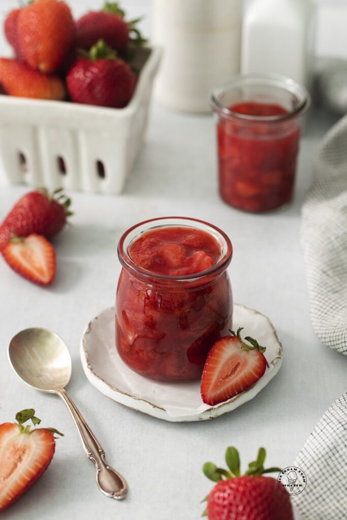 a small jar filled with homemade strawberry compote with fresh strawberries in the background.