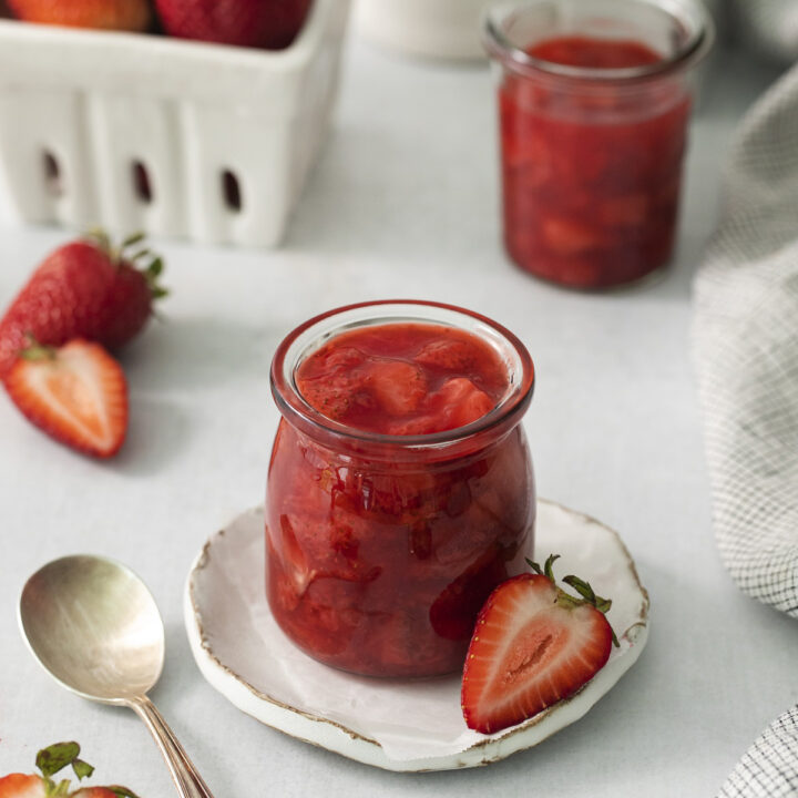 a small jar filled with homemade strawberry compote with fresh strawberries in the background.