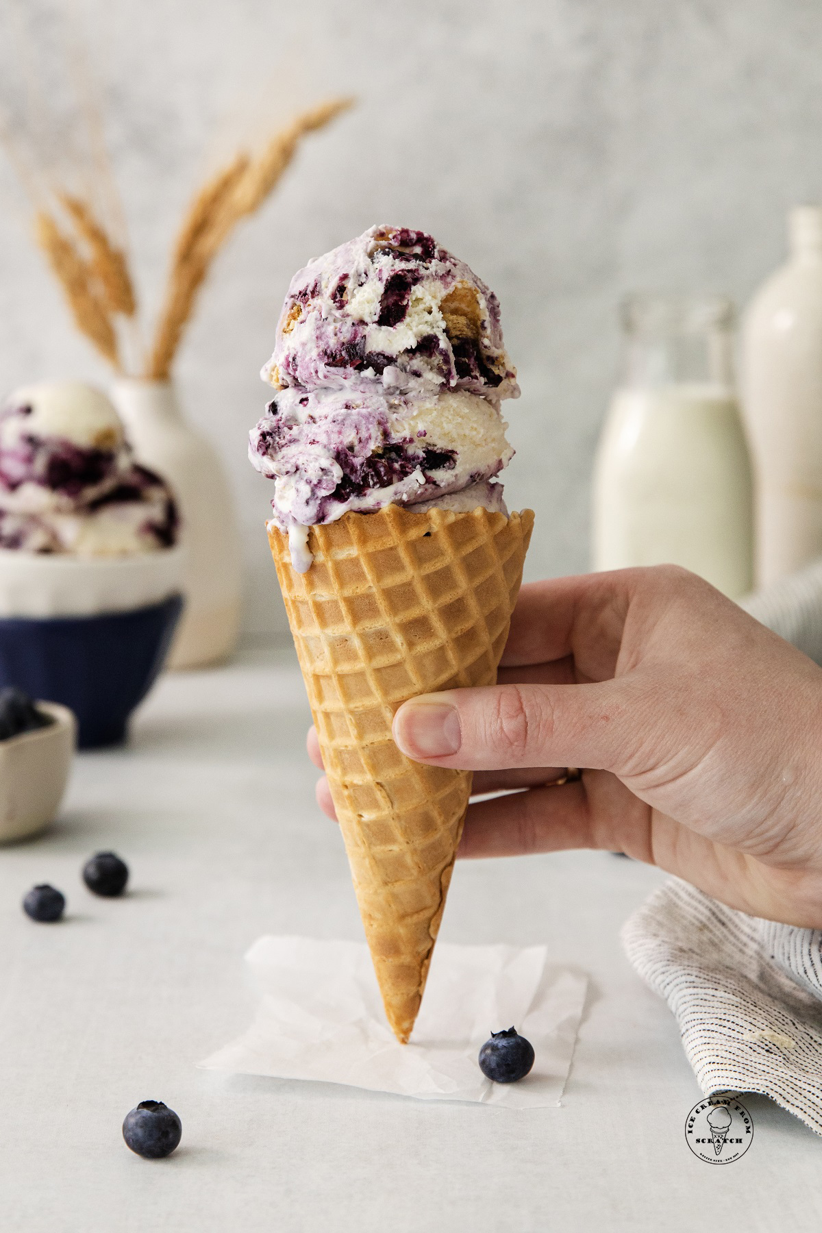scoops of blueberry cheesecake ice cream in a waffle cone, held by a hand. Bottles of milk and cream are in the background