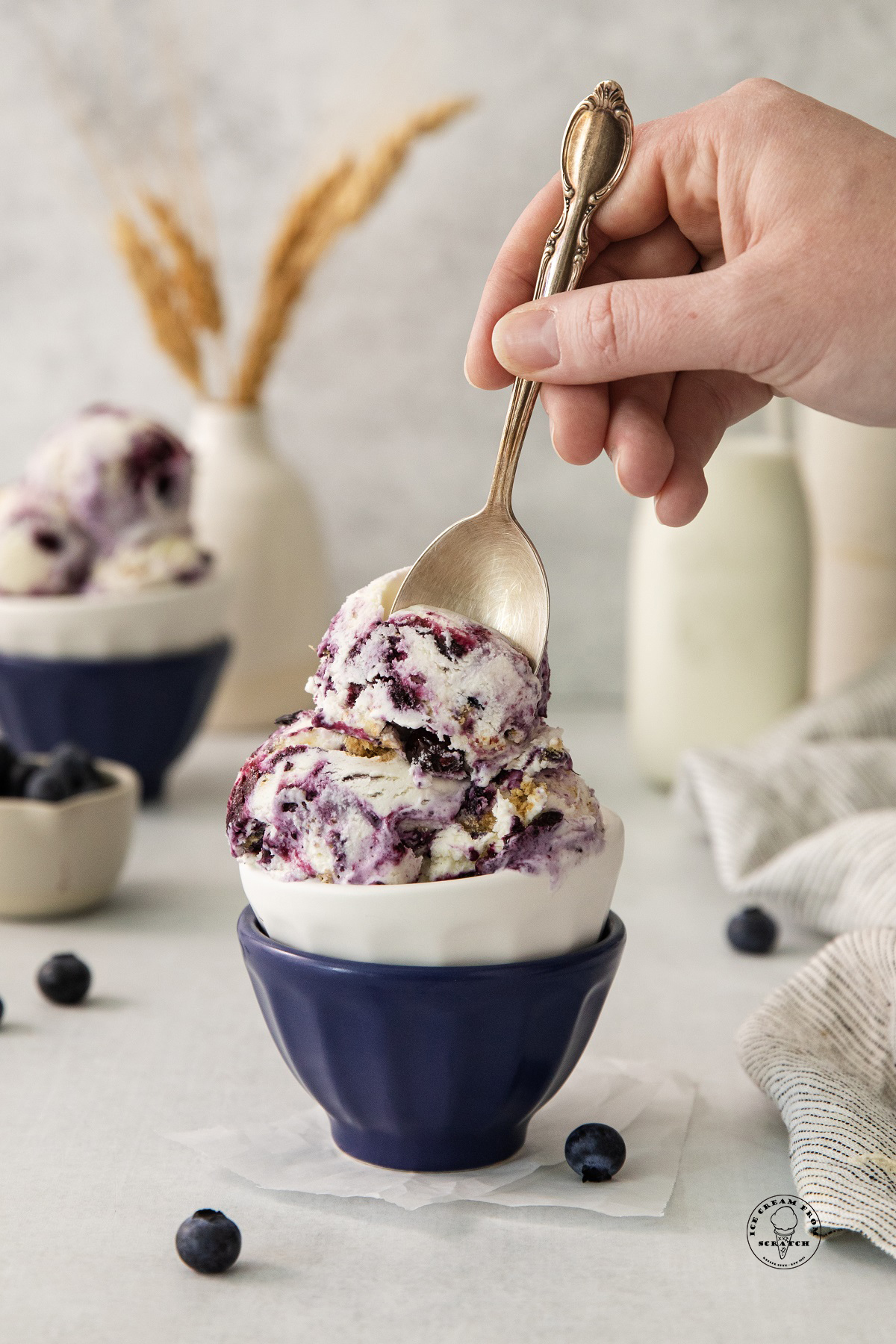 scoops of blueberry cheesecake ice cream in a small bowl, a hand is enjoying it with a spoon