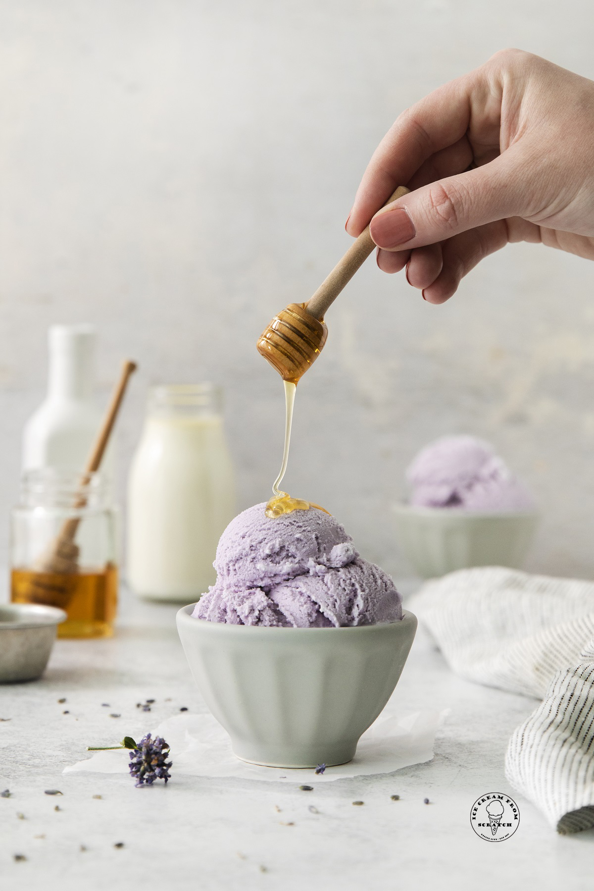 a small bowl of lavender ice cream. a hand is drizzling honey over it with a small honey dipper